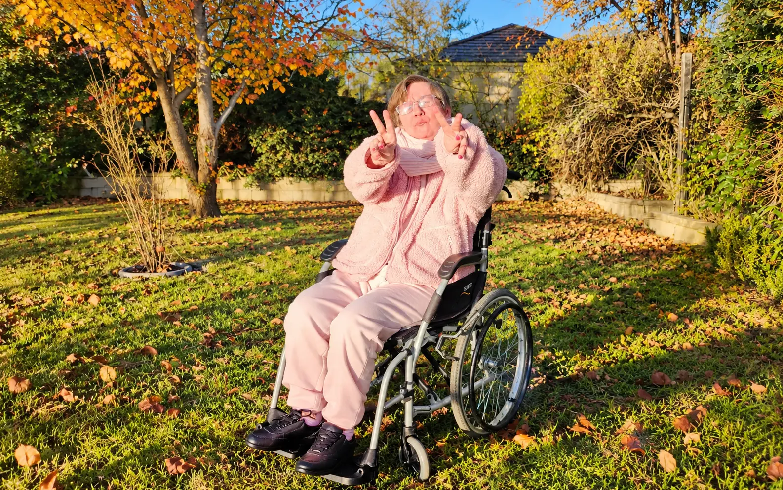 A woman in a wheelchair smiling and giving peace signs in a sunny autumn garden